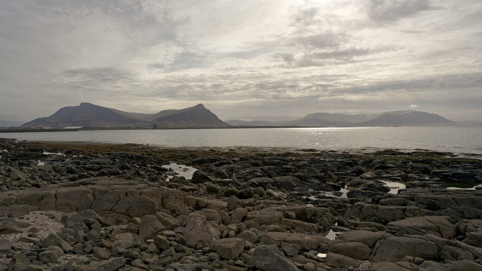 Rocky shoreline with calm sea and distant mountains under clouded sky (Akranes)