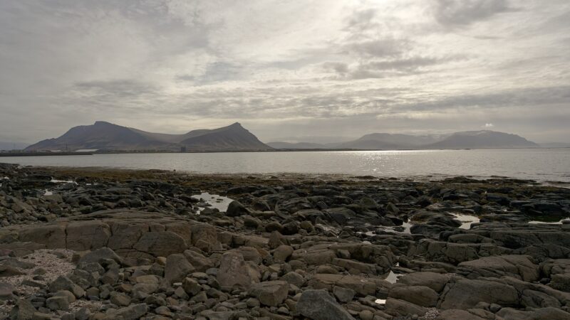 Rocky shoreline with calm sea and distant mountains under clouded sky (Akranes)