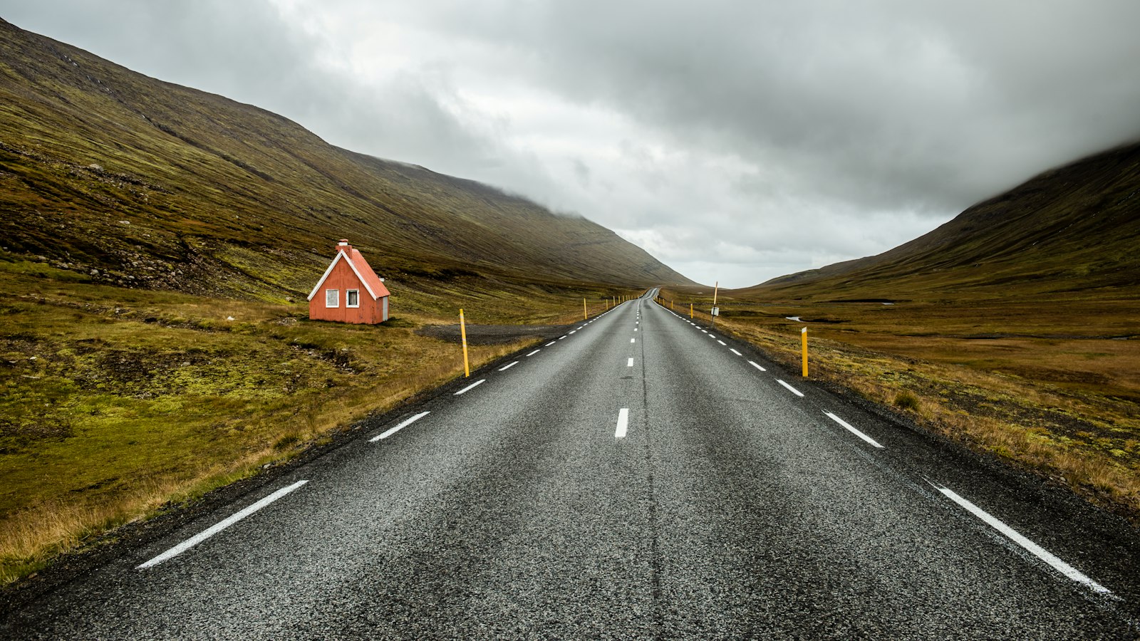 Empty straight road running between grassy hills under cloudy sky