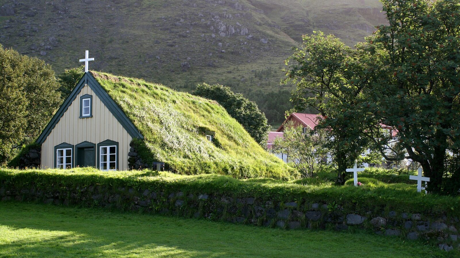 Turf-roofed church gable with a cross and trees on a grassy slope (Fagurhólsmýri)
