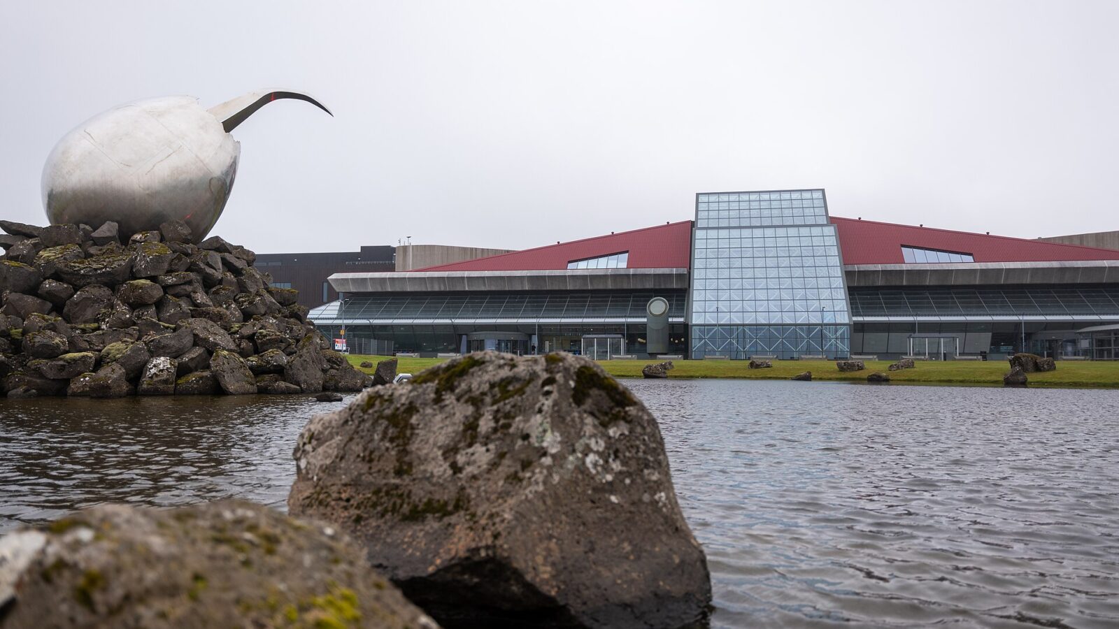 Large steel sculpture on a rocky mound beside a modern glass-front building (Keflavík International Airport)