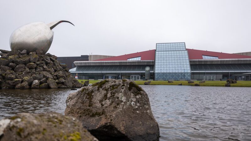 Large steel sculpture on a rocky mound beside a modern glass-front building (Keflavík International Airport)