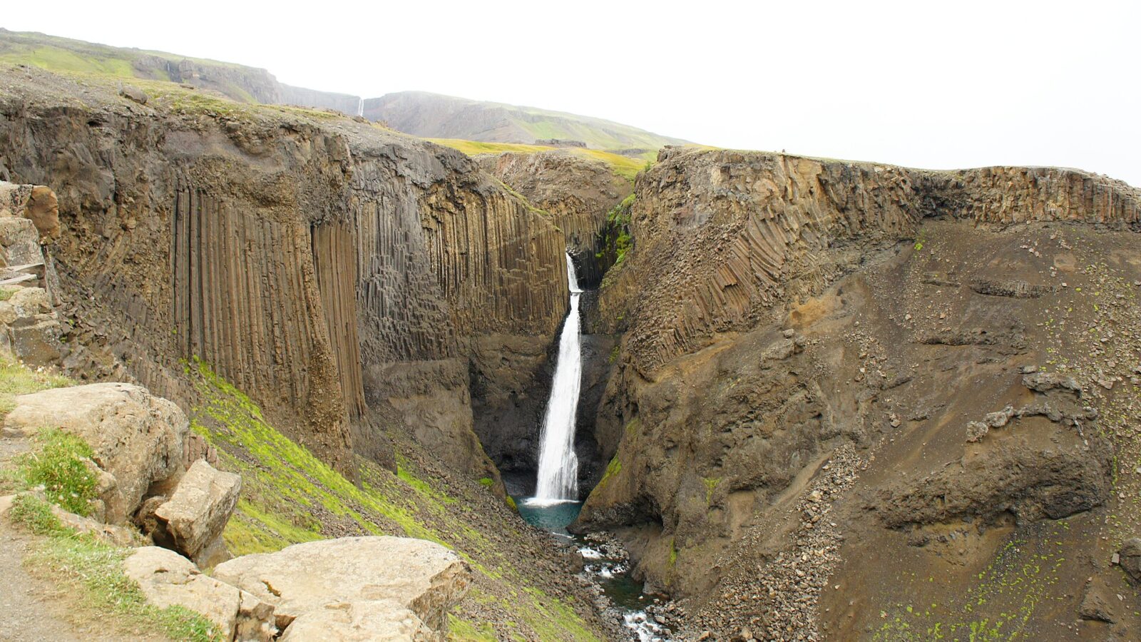 Waterfall dropping between steep basalt columns in a rocky gorge (Hjálpleysa)