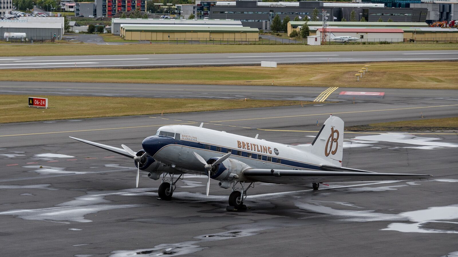 A twin‑engine vintage airliner on wet tarmac with runways and hangars beyond (Reykjavíkurflugvöllur)