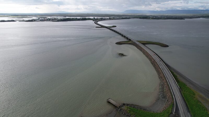 Long coastal causeway stretching toward a town and mountains. (Borgarnes)