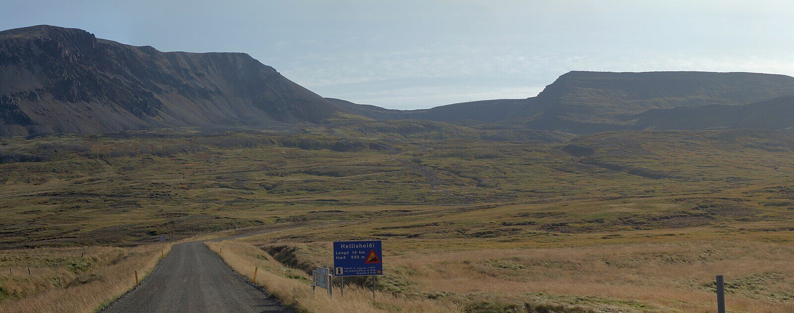Gravel road leading toward a mountain pass with a road sign (Hellisheiði)