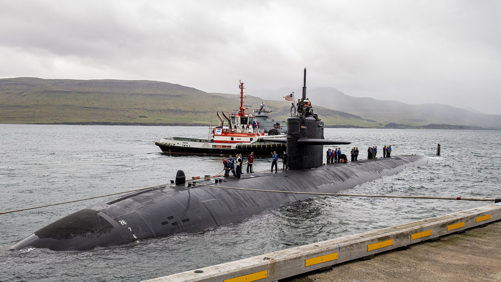 Crew members on the deck of a surfaced submarine beside a tug (Grundartangi)