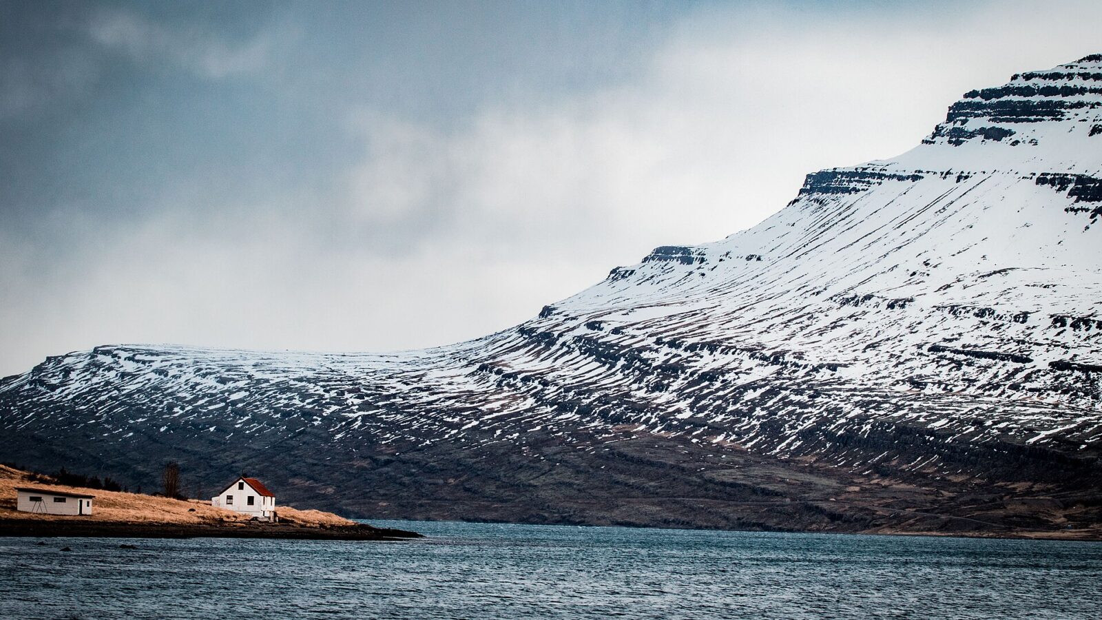 Small white house on a coastal shoreline beneath snow-covered cliffs (Reyðarfjörður)