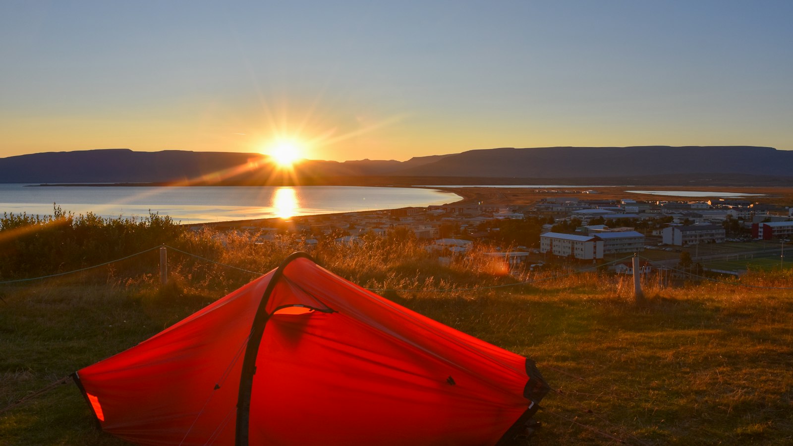 Red tent on a grassy slope with sun rising over a bay
