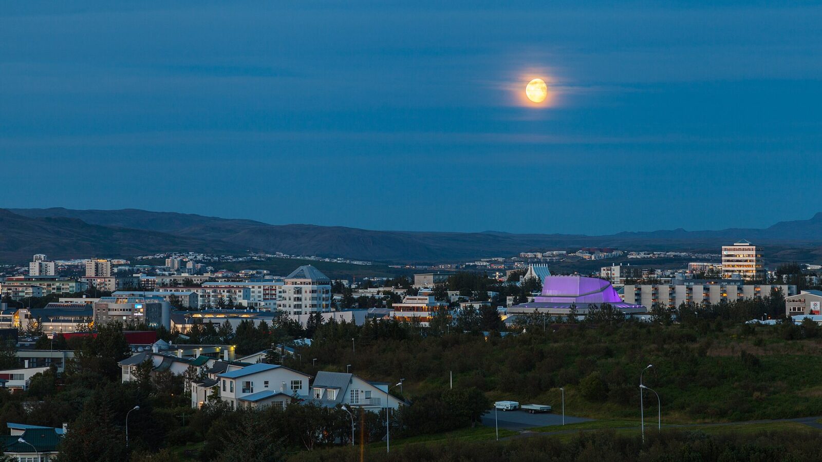 Panoramic view of a city skyline at dusk with a large moon above (Reykjavík)