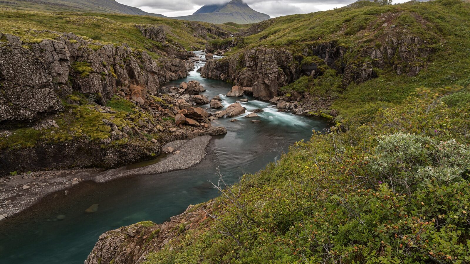 Narrow river cutting through a rocky green canyon with distant peaks (Fjarðabyggð)