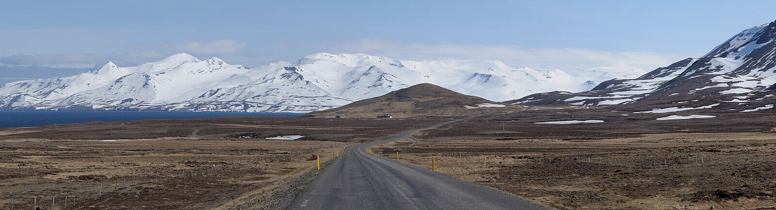 Road stretching toward a low hill with snow‑covered mountains beyond (Skagafjörður)