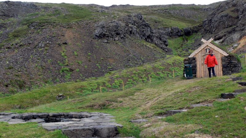 Small wooden turf hut and rocky hillside above a stone pool (Guðrúnarlaug)
