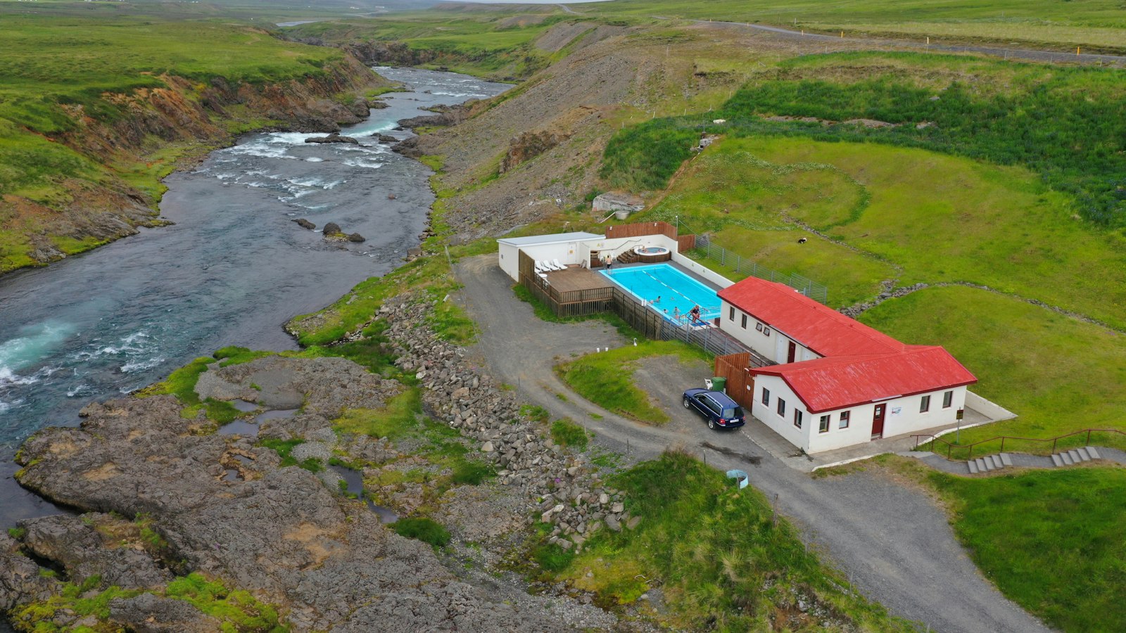 Outdoor pool and buildings beside a river and grassy hills