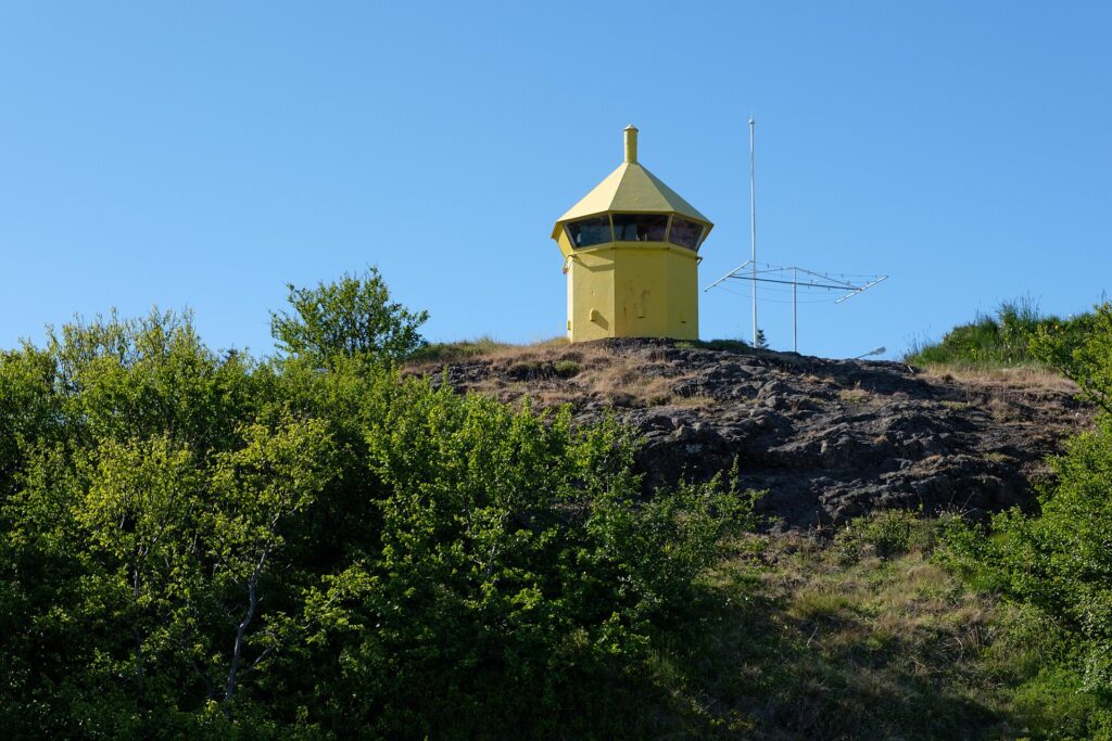 Small yellow lighthouse on a rocky hill above birch scrub (Hólmavík)