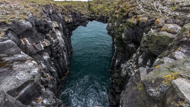 Sea arch between basalt cliffs with a person standing on the grassy rim (Snæfellsbær)