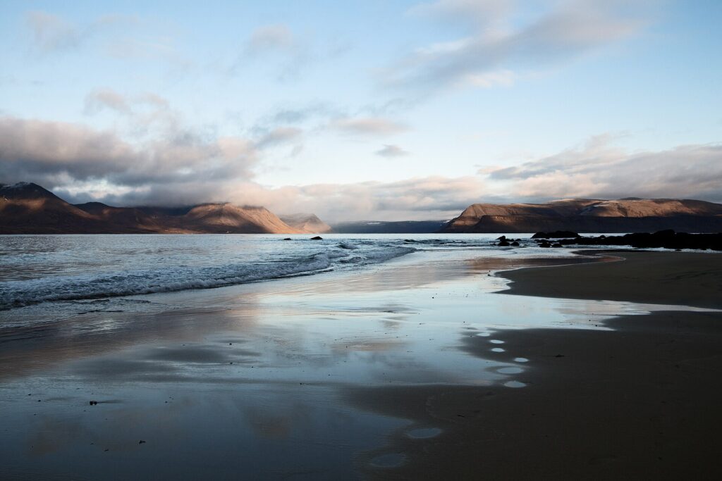 Calm beach with low waves and distant fjord mountains under a wide sky (Vesturbyggð)