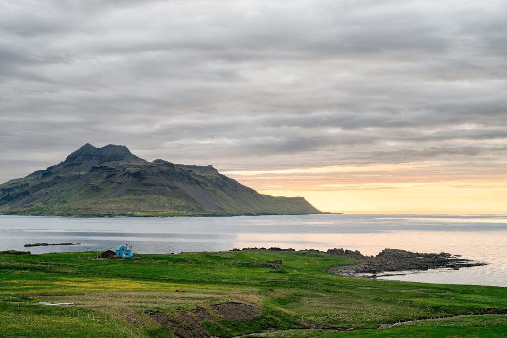 Blue farmhouse on grassy shore with mountain across the bay (Árneshreppur)