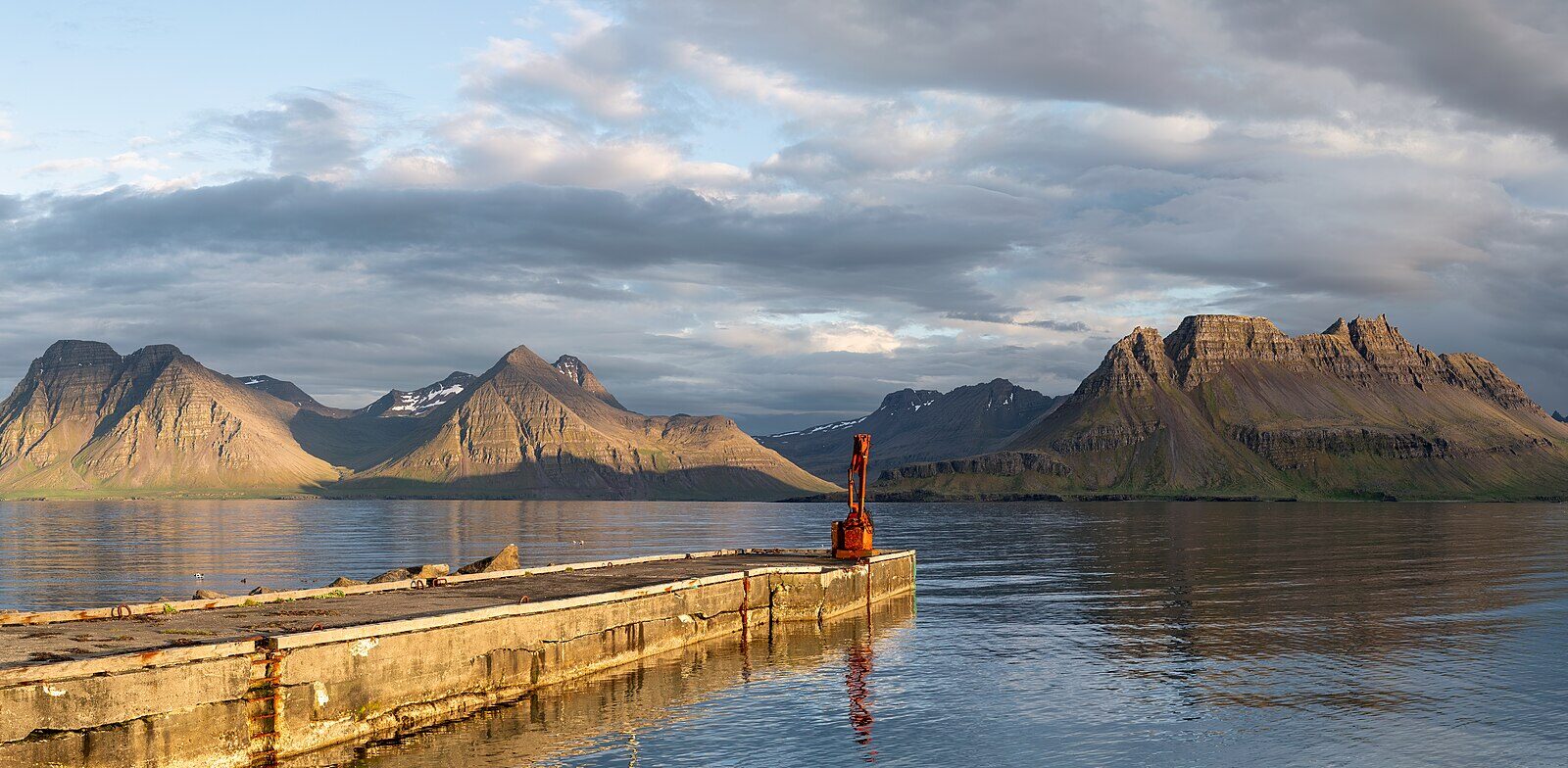 Concrete pier projecting into still water with mountains beyond (Árneshreppur)