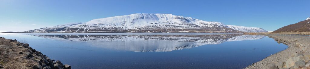 Wide view of a mountain mirrored in still water with rocky foreground (Þingeyjarsveit)