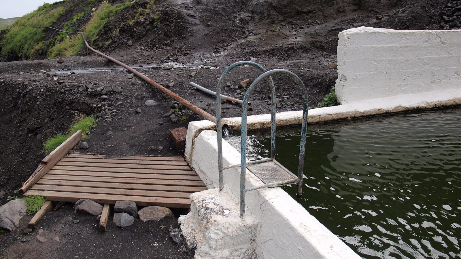 Pool ladder, concrete edge and long hot-water pipe on the hillside (Seljavallalaug)